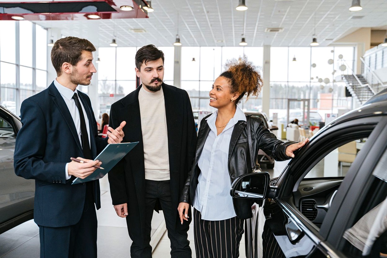A salesperson talking with a couple beside a car in a dealership showroom.